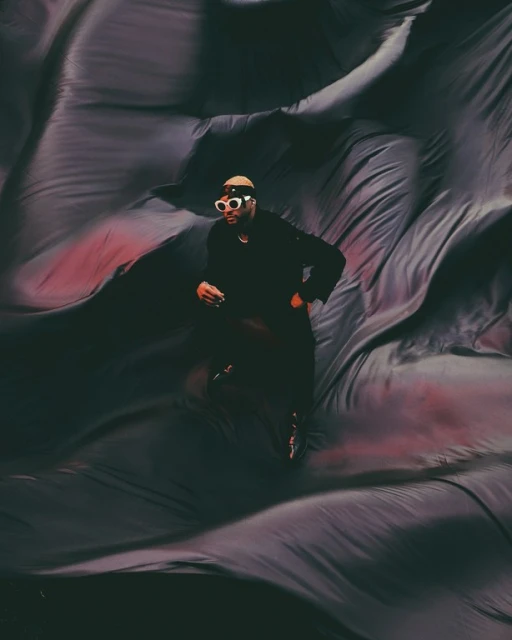 Adult male wearing sunglasses and a hat poses against a dark draped fabric background, creating a dramatic and artistic visual.