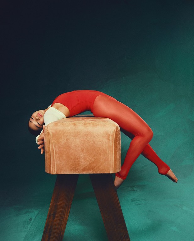 Female gymnast in red leotard and leggings reclining on a vintage brown leather pommel horse against a dark green mottled backdrop.