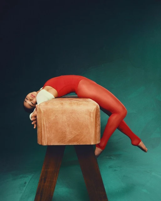 Female gymnast in red leotard and leggings reclining on a vintage brown leather pommel horse against a dark green mottled backdrop.