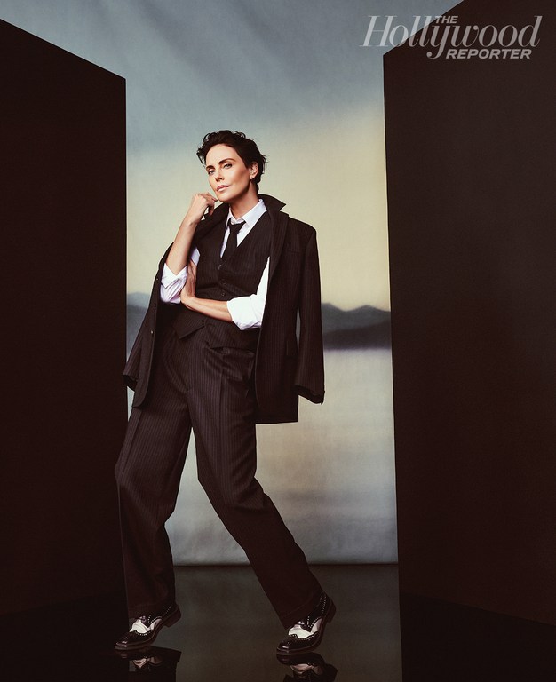 Charlize Theron in a studio portrait wearing a black suit, vest, and tie, with a white shirt and shiny black and white shoes.