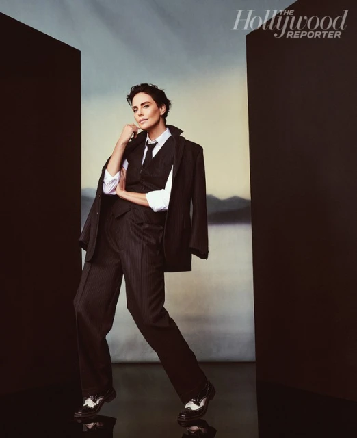 Charlize Theron in a studio portrait wearing a black suit, vest, and tie, with a white shirt and shiny black and white shoes.