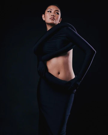 Model in black sculptural top and skirt posing against a dark background, photographed by Chrisean Rose.
