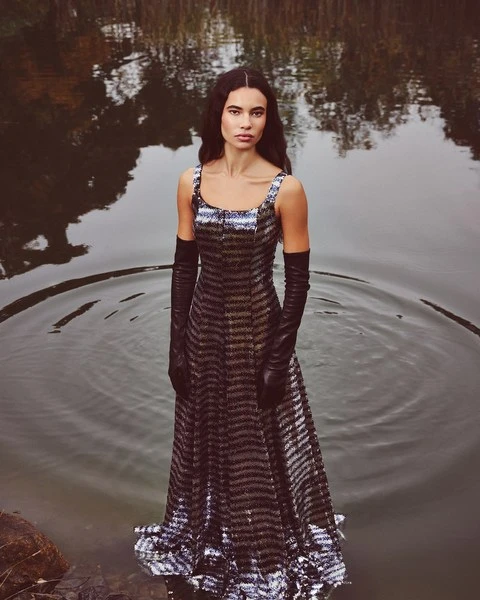 A woman in a dress stands knee-deep in a lake surrounded by trees and foliage, creating a nature and fashion editorial shot.