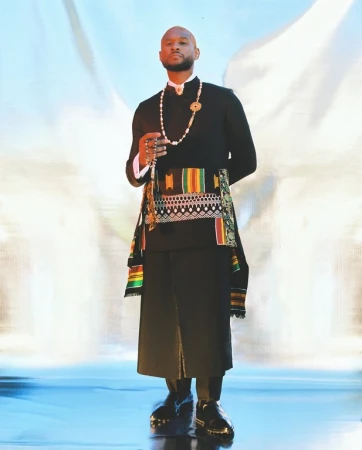 Black man wearing a black tunic, patterned sash, beaded necklace and holding prayer beads against a bright backdrop.