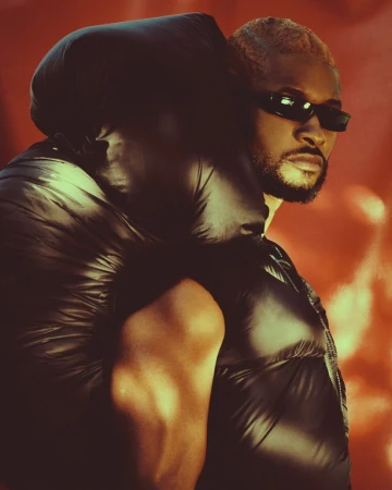 Black man wearing sunglasses and a black puffer vest against a warm orange background, photographed by Chrisean Rose.