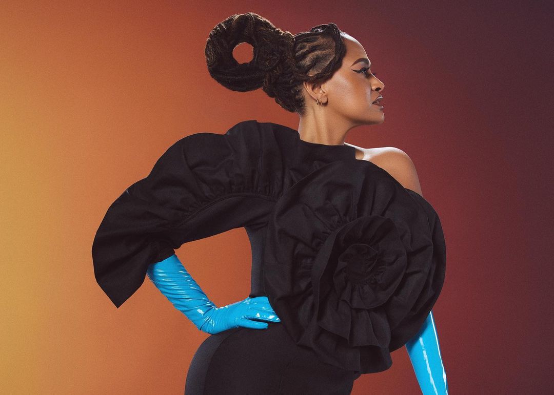 Portrait of an African-American woman in a black dress with exaggerated ruffles and blue latex gloves against an orange background.