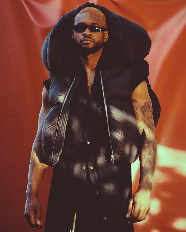 Black man with dyed hair wearing dark sunglasses and a black puffer vest against a terracotta background