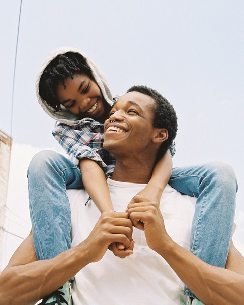 An African-American father giving his son a piggyback ride, both are smiling against a bright outdoor urban background.