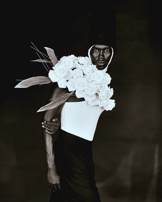 Portrait of a Black woman in a white top adorned with white roses and leaves, set against a dark backdrop in a studio photoshoot.