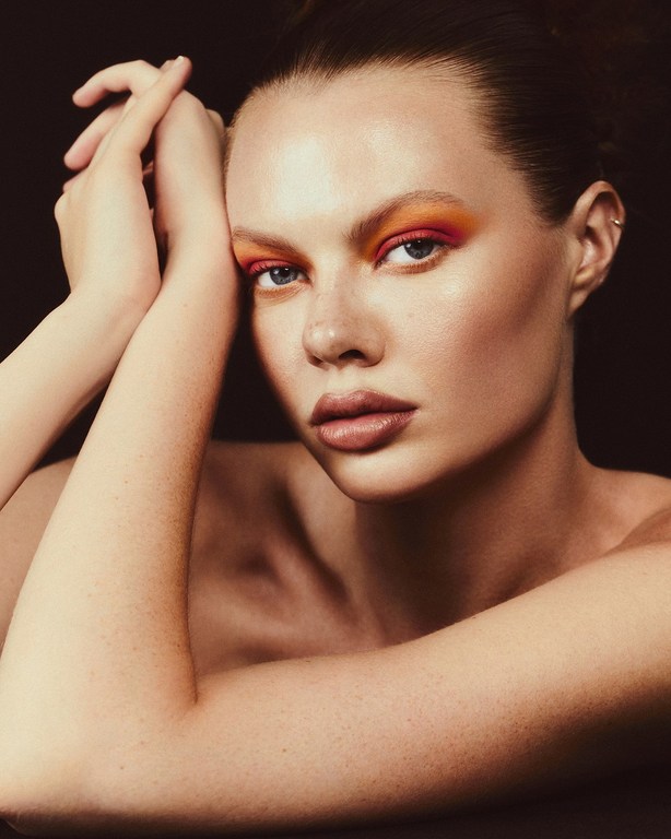 Close-up beauty shot of a fair-skinned woman with orange eyeshadow and dark lipstick, posing with her hands near her face, against black.
