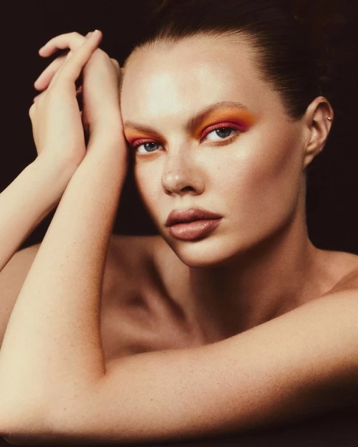 Close-up beauty shot of a fair-skinned woman with orange eyeshadow and dark lipstick, posing with her hands near her face, against black.