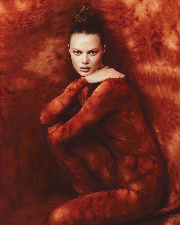 Portrait of a woman in a long-sleeved jumpsuit crouching against a mottled red background, wearing a floral hairpiece in an indoor environment.