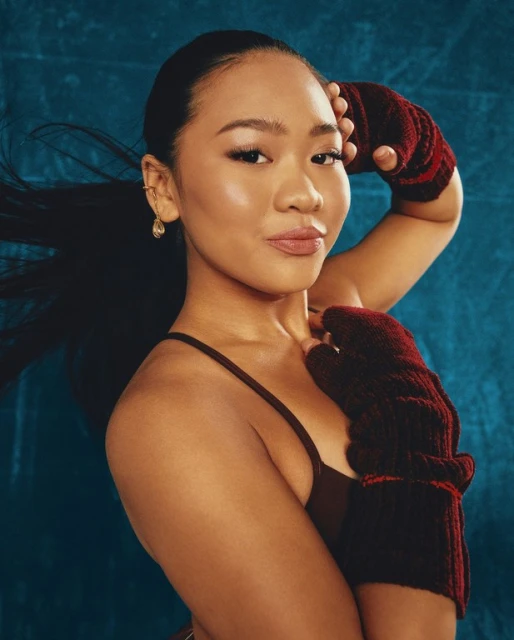 Portrait of a smiling woman with long dark hair, wearing a brown top and maroon knit fingerless gloves, against a teal background.