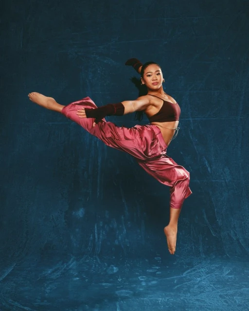 Athletic young woman with dark hair in a high ponytail poses in a dynamic dance leap wearing flowing pants and a dark top against a blue backdrop.