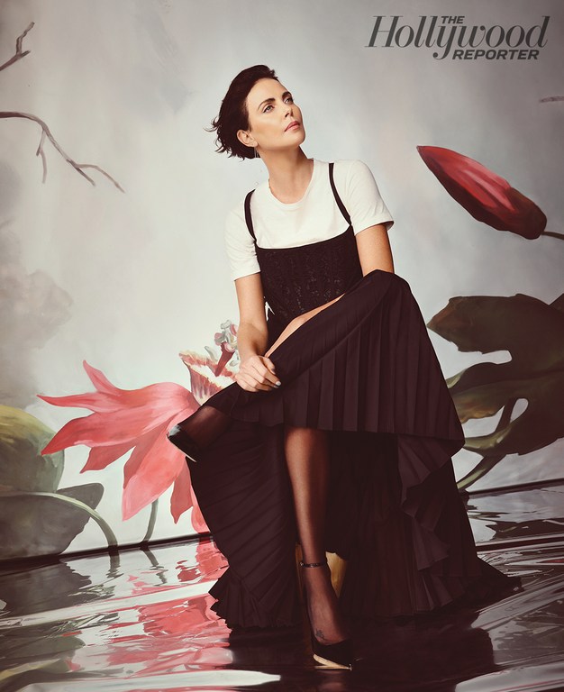 Portrait of Charlize Theron in a black dress over a white t-shirt sitting in front of a floral studio backdrop.