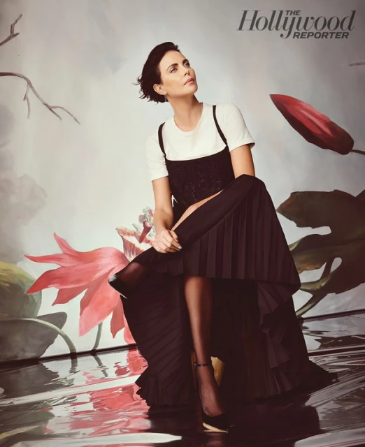 Portrait of Charlize Theron in a black dress over a white t-shirt sitting in front of a floral studio backdrop.
