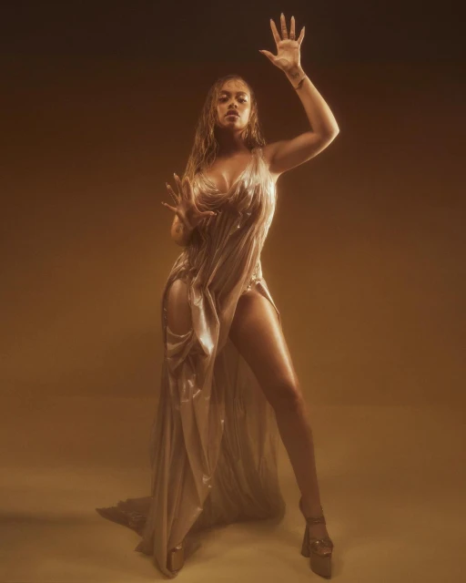 Woman in shimmering gown strikes pose in warm light. Full shot shows floor-length dress, raised hand, and platform heels against soft gold backdrop.