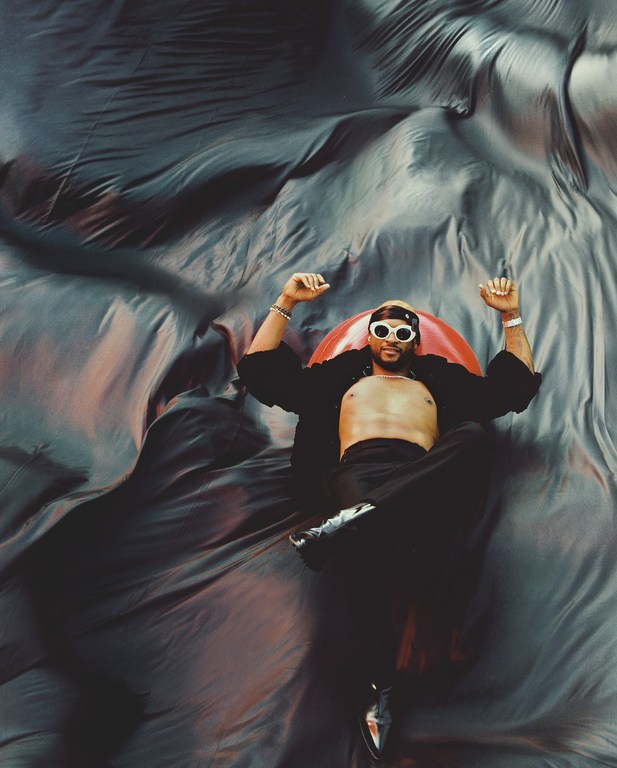 Man lying on a large piece of fabric in a red inflatable tube, wearing sunglasses, an open black shirt, pants, and dress shoes.