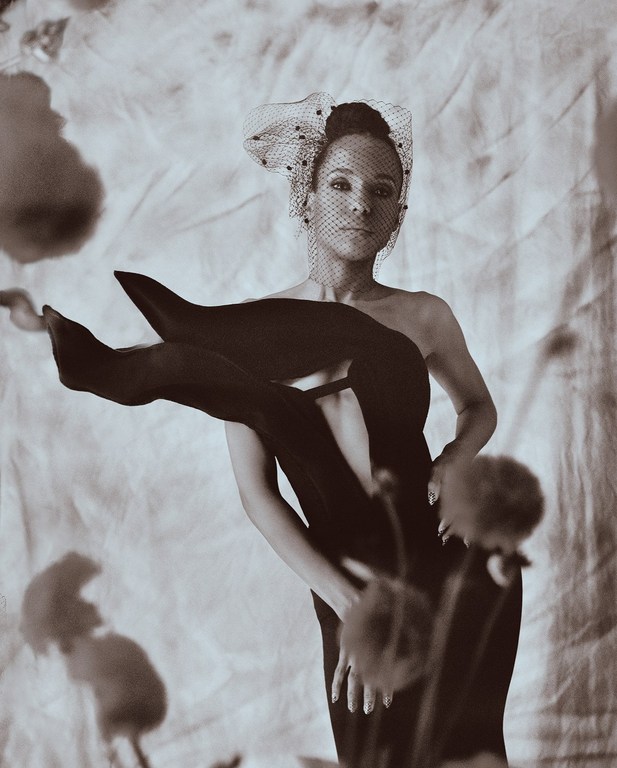 Elegant woman in a black dress and veil poses in a studio with floral accents against a textured backdrop, monochrome visual style.