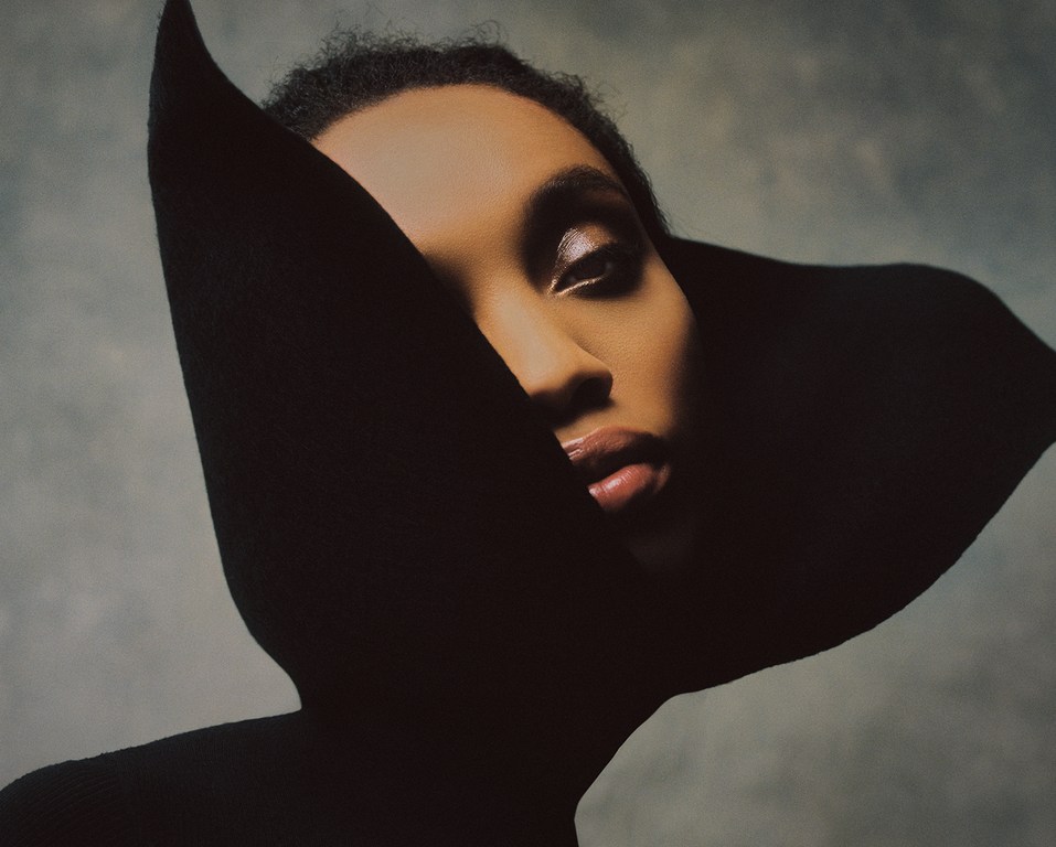 Striking portrait of a woman with dark hair and glamorous makeup partially obscured by a large, dark, sculptural element, set against a soft grey backdrop.