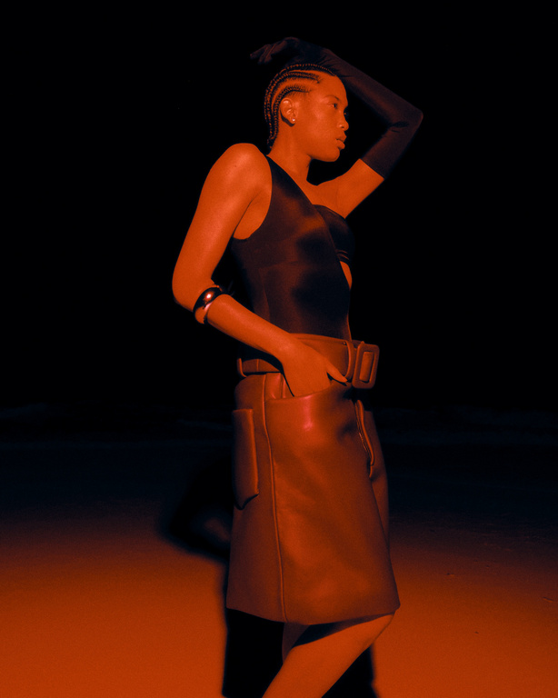 Stylish woman in a dark setting posing in a one-shoulder top, belted skirt, and long gloves with a glowing, warm-toned gel light.