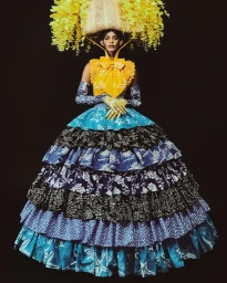 Model wearing a structured yellow top, tiered blue patterned skirt, and a large basket headdress adorned with yellow flowers.