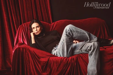 Charlize Theron reclining on a draped red velvet sofa wearing a black turtleneck and blue jeans