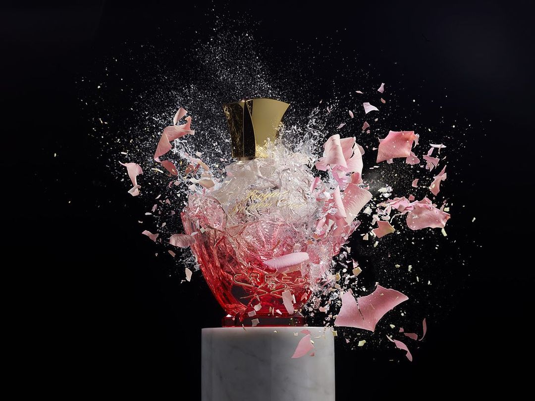 Artistic image of a shattered perfume bottle on a marble stand against a black background. Sharp shards and liquid are suspended in air.