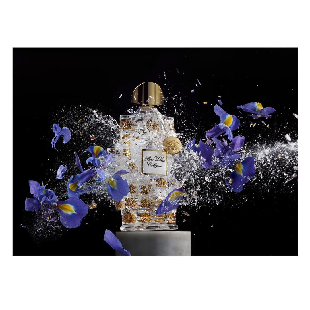 Perfume bottle surrounded by blue iris flowers and water droplets on a gray pedestal against a black background in studio lighting.