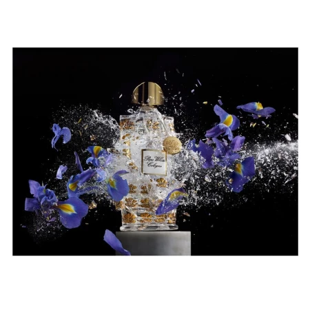 Perfume bottle surrounded by blue iris flowers and water droplets on a gray pedestal against a black background in studio lighting.