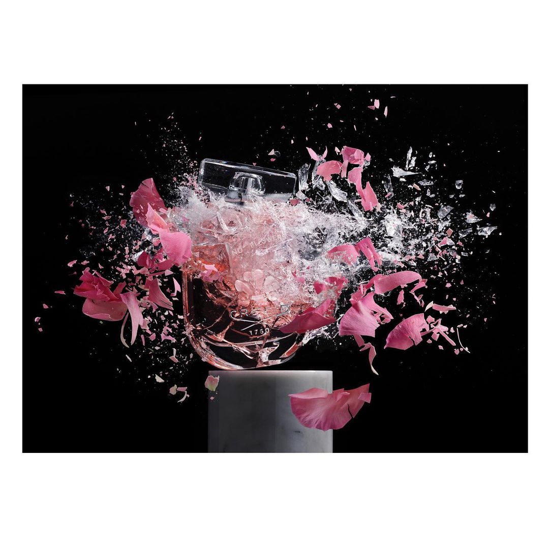 Still life of a perfume bottle on a stand, surrounded by pink flower petals and shattering glass against a stark black backdrop.