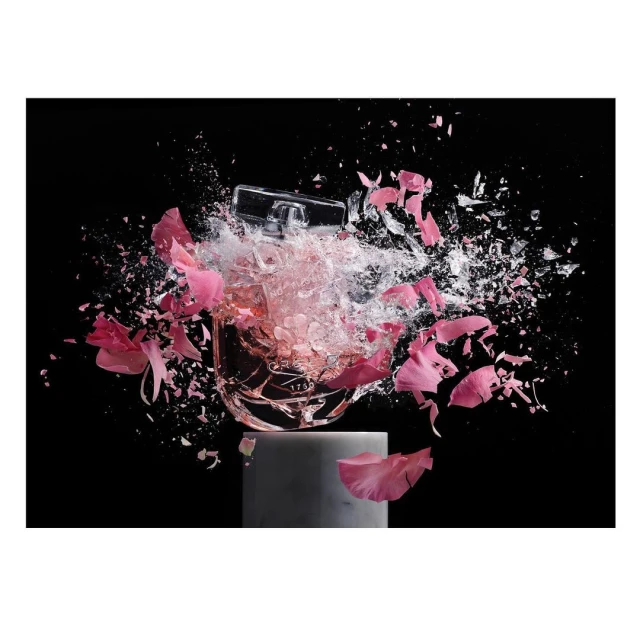 Still life of a perfume bottle on a stand, surrounded by pink flower petals and shattering glass against a stark black backdrop.