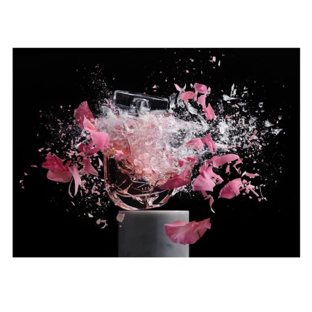 Still life of a perfume bottle on a stand, surrounded by pink flower petals and shattering glass against a stark black backdrop.