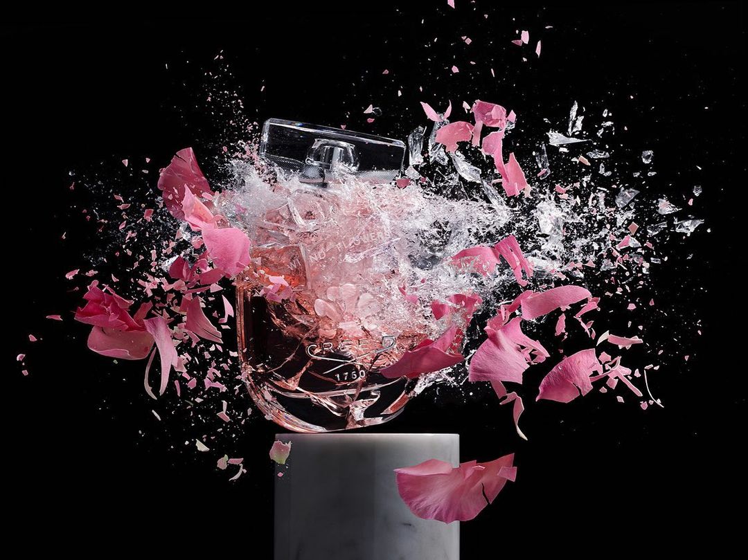 Perfume bottle explodes on marble plinth amidst shattering glass and scattered pink flower petals against a solid black backdrop.