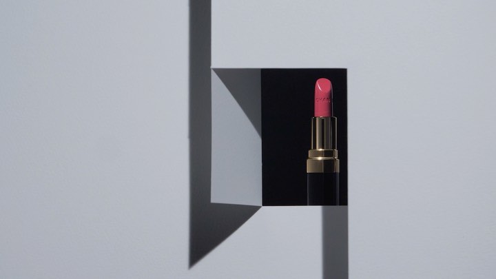 Close-up of pink lipstick with gold accents against a geometric black, white, and gray block design in a beauty product still life.