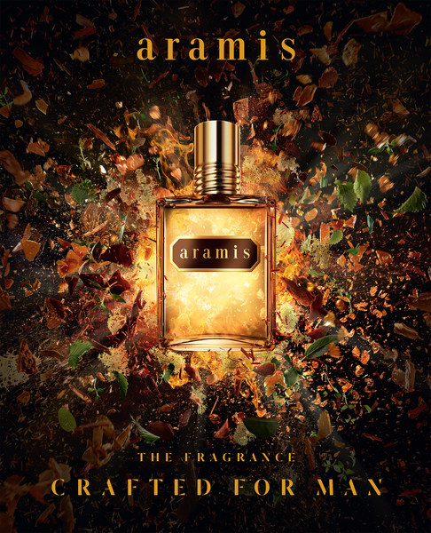 A crystal Aramis perfume bottle is centered, with leaves and wood fragments swirling in a dynamic composition against a black background.