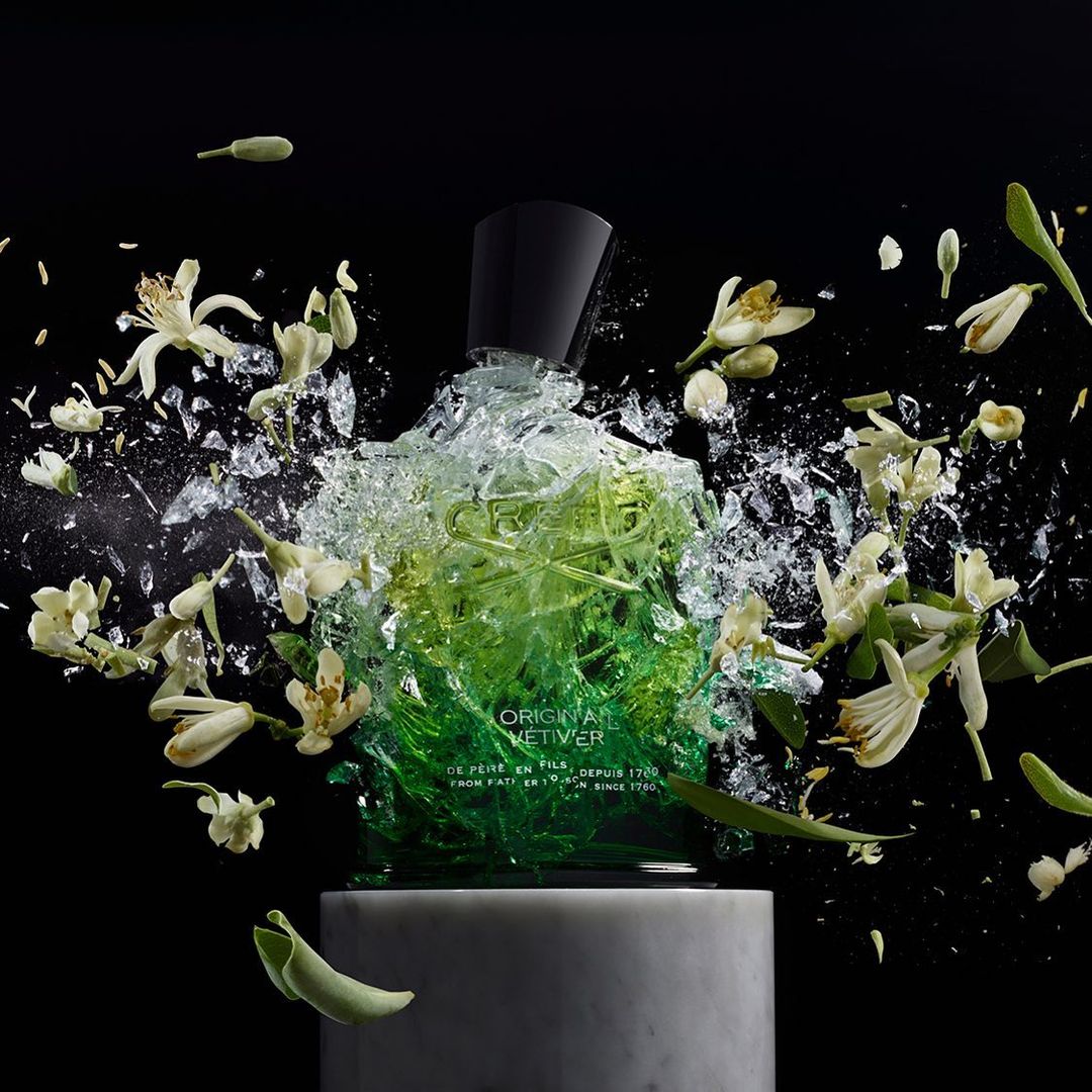 A glass bottle of green and clear Creed Original Vetiver fragrance on a marble pedestal with white flowers against black background.