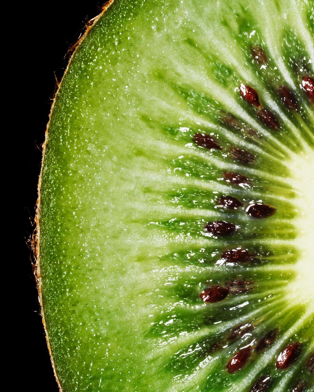 Close-up of a vibrant green kiwi fruit slice featuring tiny dark seeds and a pale yellow center against a stark black backdrop.