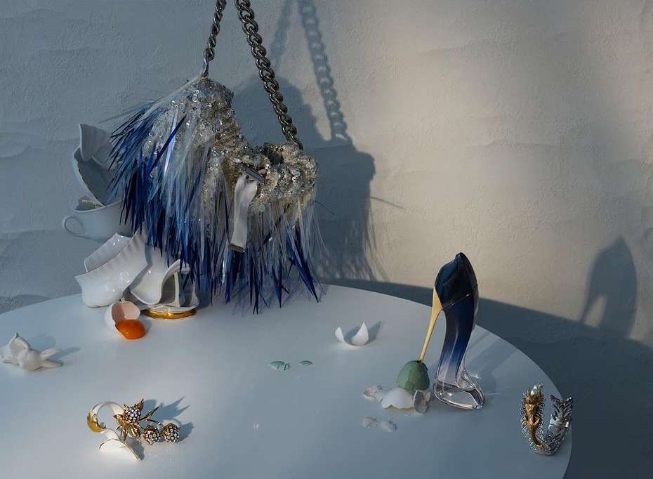 Still life featuring a handbag with blue fringe, a pair of high heels, eggshell, and shattered china on a white table against a white wall.