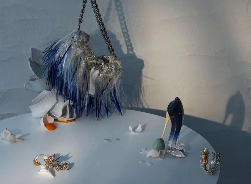 Still life featuring a handbag with blue fringe, a pair of high heels, eggshell, and shattered china on a white table against a white wall.