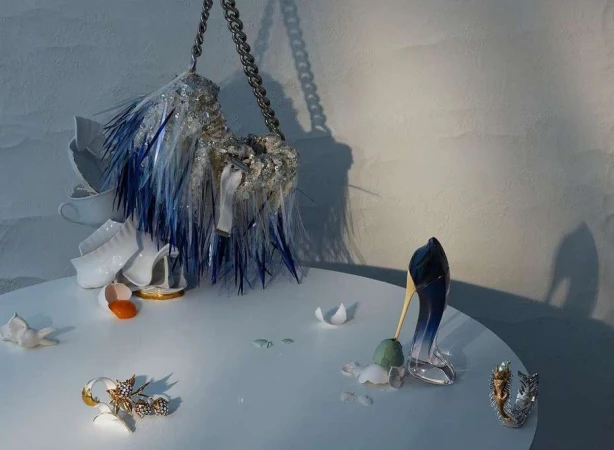 Still life featuring a handbag with blue fringe, a pair of high heels, eggshell, and shattered china on a white table against a white wall.