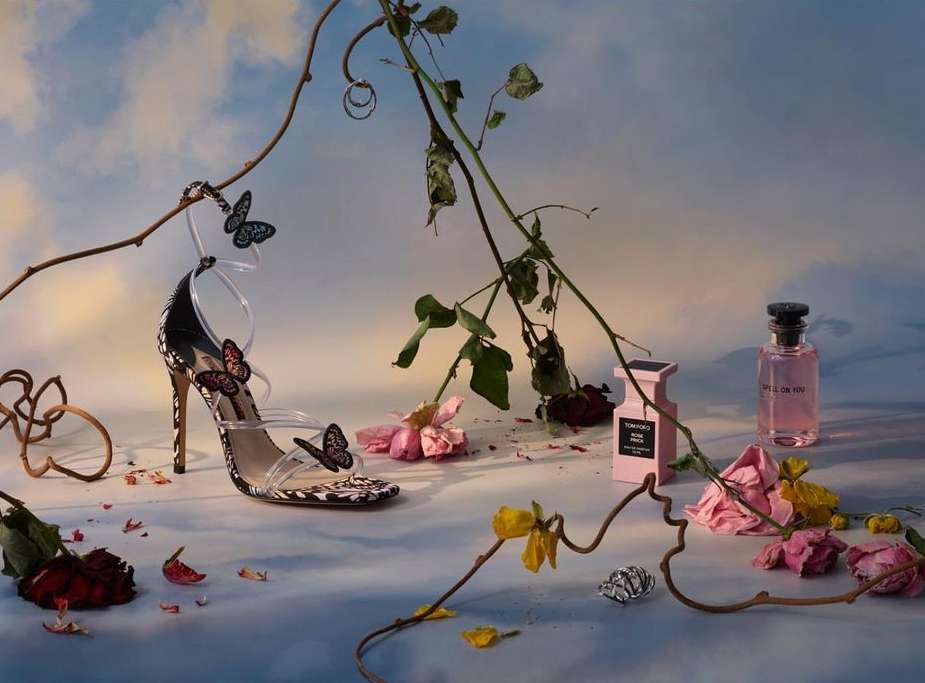 High-heeled shoe with butterfly detail beside two perfume bottles and scattered roses and flower petals on a light blue background.