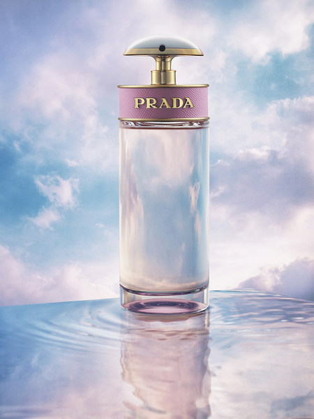 Prada fragrance bottle on a reflective surface with a dreamy sky background, showcasing its elegant design and soft, ethereal color palette.