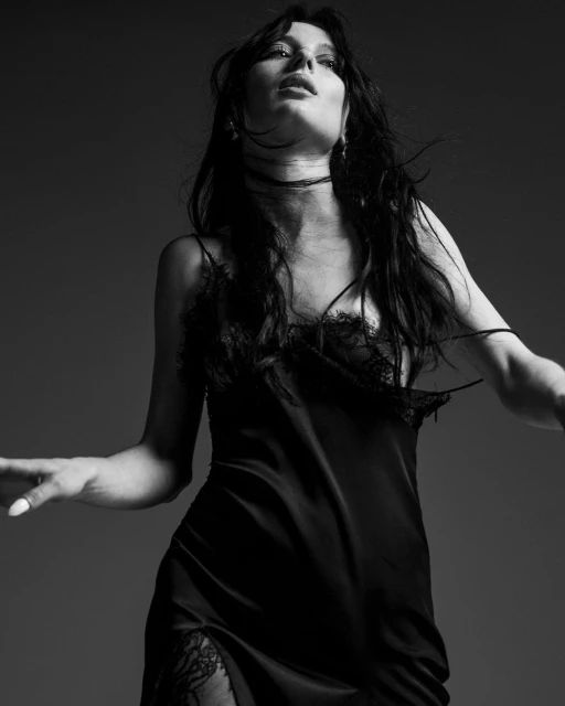 Monochrome portrait of a woman in a black lace-trimmed slip dress, looking upward with a soft expression against a grey backdrop.