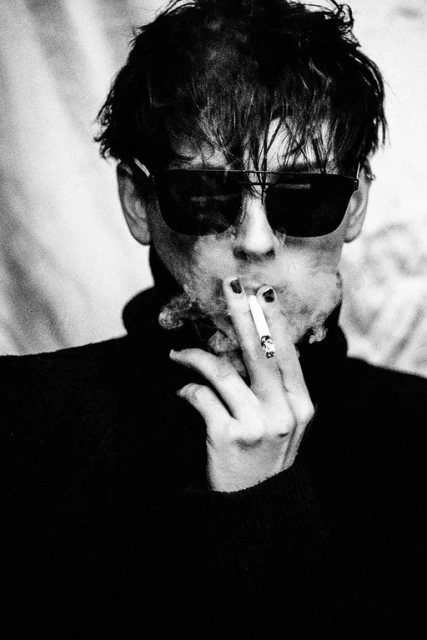 Black and white portrait of a young adult smoking a cigarette, wearing dark sunglasses and a black turtleneck sweater. Smoke is rising.