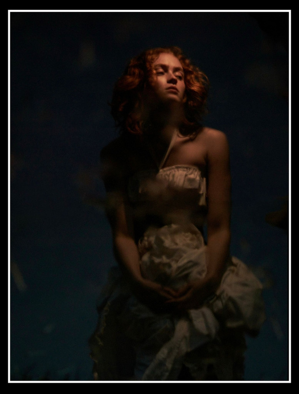 A woman with red curly hair in a strapless dress with a dark blue background is softly lit in a studio-style portrait, creating a moody image.