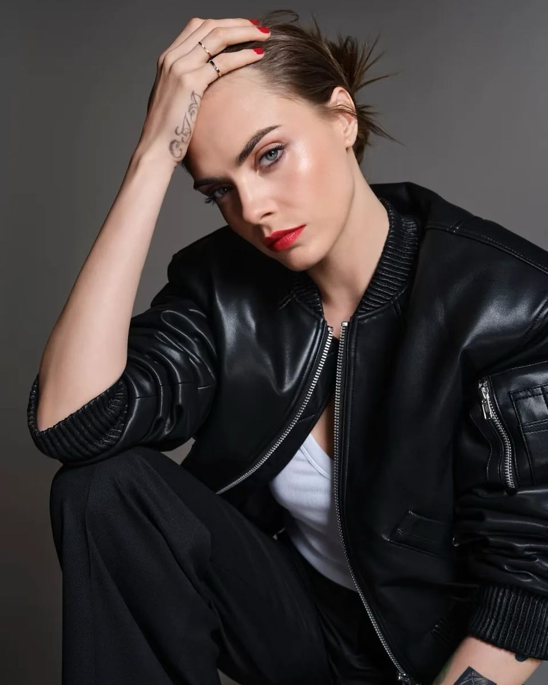 Cara Delevingne in a black leather bomber jacket posing against a neutral grey studio background by photographer Damon Baker