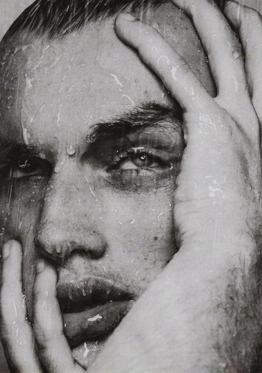 Close-up monochrome portrait of a man with wet hair and water droplets on his face, holding his head with both hands.
