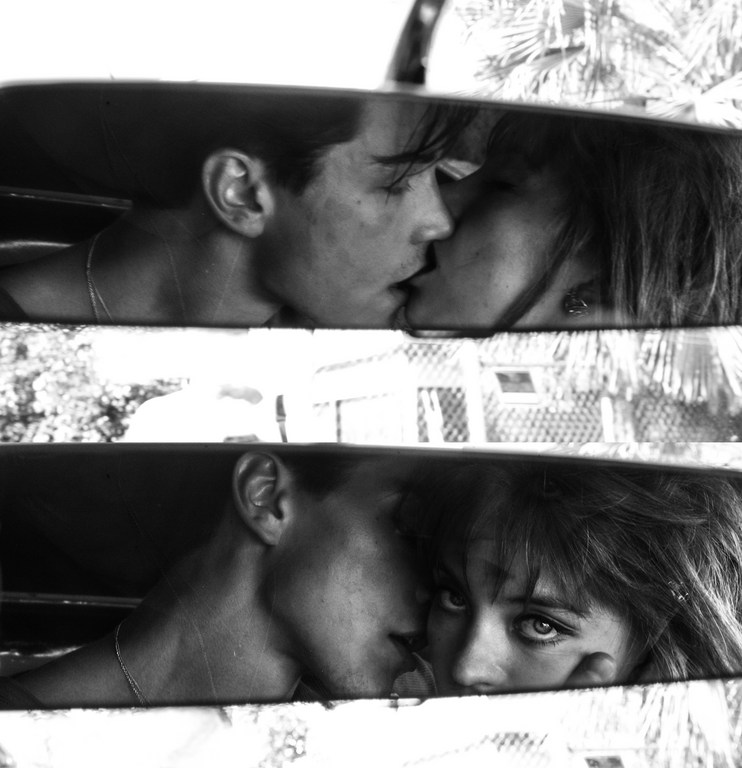 Black and white shot of couple reflected in car mirror, kissing intimately. Outdoor setting with building and foliage visible in the background.