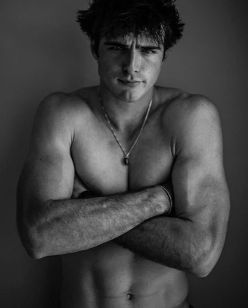 Portrait of a shirtless man with dark hair, arms crossed and wearing a pendant necklace against a dark background
